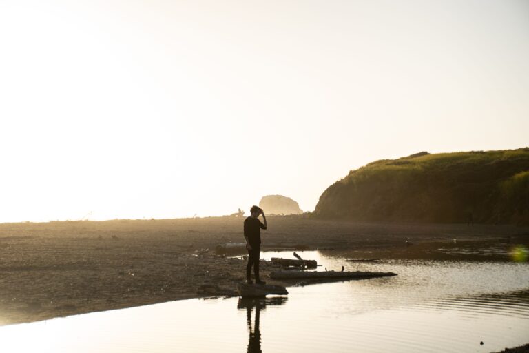 Silhouette of a man in black outfit standing on a coastal shore during sunset, capturing the natural beauty.
